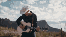 a man in a cowboy hat is playing an acoustic guitar in a field with mountains in the background .