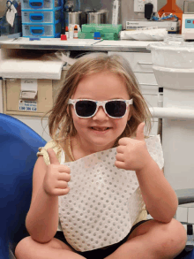 a little girl wearing sunglasses and a bib gives a thumbs up