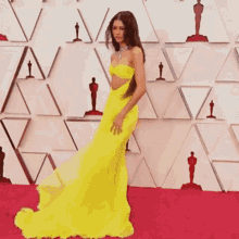 a woman in a yellow dress is standing on a red carpet in front of a wall with oscar statues .