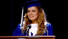 a woman in a graduation cap and gown stands at a podium giving a speech that says life is short .