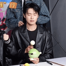 a man in a black leather jacket is sitting at a table holding a green frog toy .