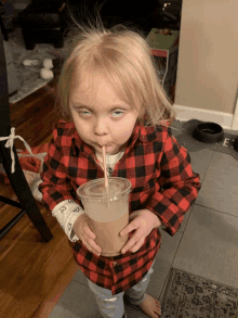 a little girl in a plaid shirt drinks from a cup with a straw