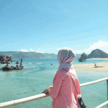 a woman in a pink shirt and polka dot head scarf stands on a railing overlooking the ocean