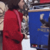 a woman in a red coat is standing next to a blue trash can with the word police on it