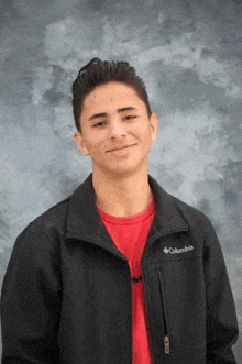 a young man wearing a black columbia jacket and a red shirt is smiling .