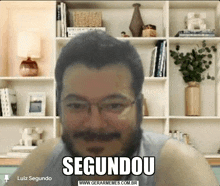 a man with glasses and a beard is sitting in front of a bookshelf with the word segundou on his face .