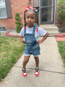 a little girl wearing overalls and red and white sneakers stands on a sidewalk