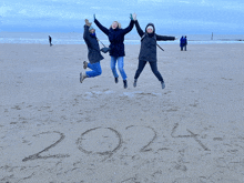 three people are jumping in the air on a beach with the year 2024 written in the sand