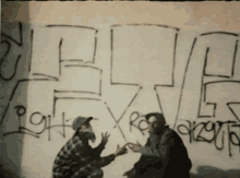 two men squatting in front of a wall that has graffiti on it including the letters lg