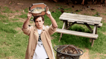 a man in a trench coat holds up a plaque with a fish on it in front of a picnic table