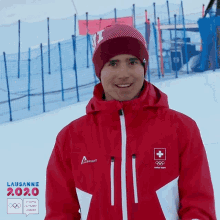 a man wearing a red jacket and a beanie is smiling in front of a sign that says lausanne 2.0.0