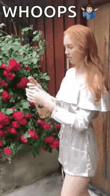 a woman in a white dress is standing in front of a bush of red roses and looking at her phone