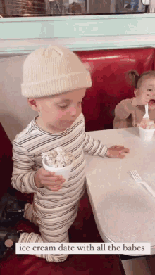 a little boy is holding a cup of ice cream while sitting at a table with two other children .