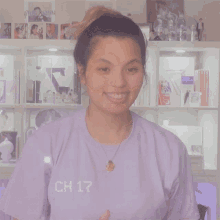 a woman wearing a purple shirt and necklace smiles in front of a shelf with a picture of a man on it