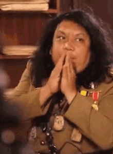 a woman in a military uniform is sitting in front of a bookshelf with her hands folded in front of her face .