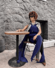 a woman in a blue dress sits at a table in front of a brick wall