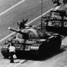 a black and white photo of a man standing in front of a row of tanks on a street .