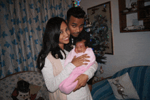 a man and woman holding a baby in front of a christmas tree in a living room