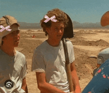 two young men are standing next to each other in the desert . one of the men is wearing a pink bow in his hair .