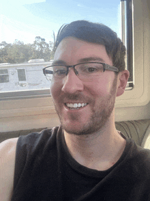 a man wearing glasses and a black tank top smiles in front of a window