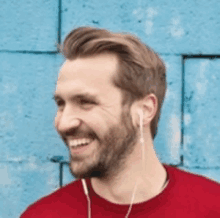 a man with a beard is wearing ear buds and smiling in front of a blue wall .