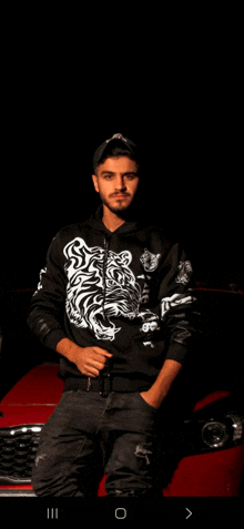 a man wearing a black jacket with a tiger on it leans on a red car