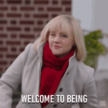 a woman in a white coat and red scarf is standing in front of a brick wall and says welcome to being .