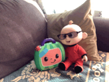 a stuffed baby wearing sunglasses sits on a couch next to a stuffed watermelon