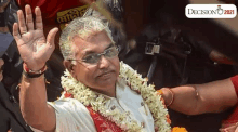 a man wearing glasses and a garland of flowers is waving his hand in the air .