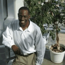 a man in a white shirt and khaki pants is sitting next to a potted plant .