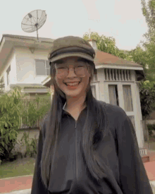 a woman wearing a hat and glasses is smiling in front of a house with a satellite dish on the roof .