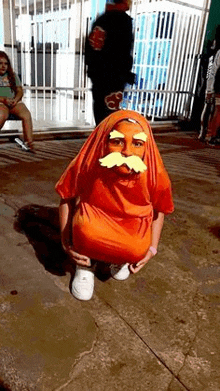 a young boy in an orange costume with a mustache is squatting down on the ground .