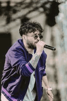 a man with a beard is singing into a microphone while wearing sunglasses and a purple jacket .