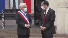 two men wearing face masks are standing next to each other in front of a sign that says " mediabanco "