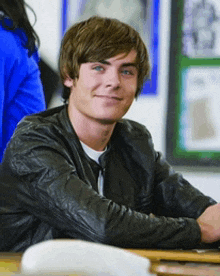 a young man wearing a leather jacket sits at a desk