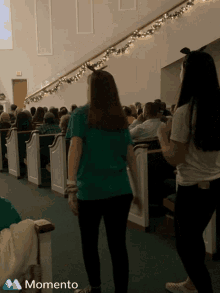 a woman in a green shirt stands in front of a crowd of people in a church with a momento logo in the corner