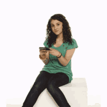 a woman in a green shirt is sitting on a white cube looking at her phone