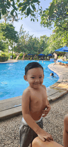 a shirtless little boy stands in front of a large swimming pool