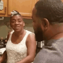 a man and a woman are standing in a kitchen talking to each other . the woman is wearing a white tank top .