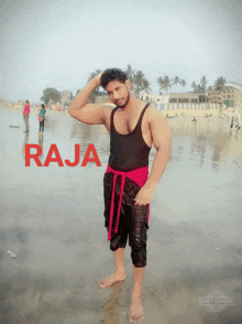 a man in a black tank top and red shorts stands on a beach with the word raja in red