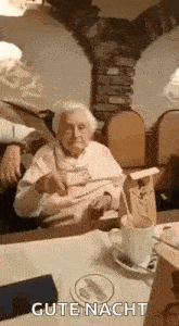 an elderly woman is sitting at a table with a cup of coffee and a spoon in her mouth .