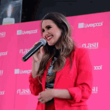 a woman in a red jacket is singing into a microphone in front of a pink wall that says liverpool