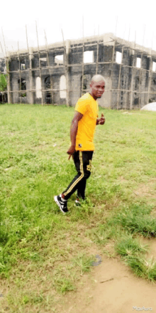 a man in a yellow shirt and black pants is standing in a grassy field in front of a building under construction