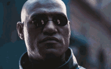 a bald man wearing sunglasses and a leather jacket looks at the camera