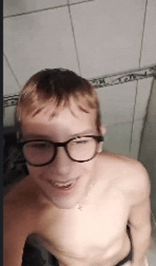 a shirtless young man wearing glasses is smiling while taking a selfie in a bathroom .