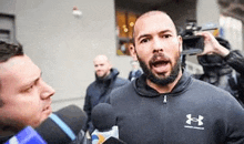 a man with a beard is talking into a microphone while a cameraman takes a picture of him .