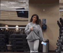 a woman in a grey sweatshirt is standing in a gym next to a trash can and a stack of dumbbells