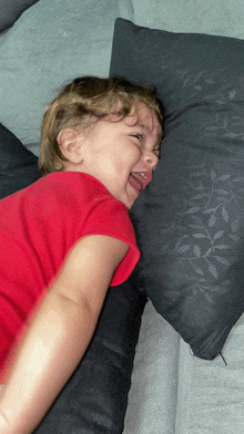 a little boy in a red shirt is laying on a black pillow with his tongue out