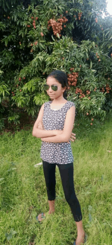 a girl wearing sunglasses is standing in the grass in front of a tree with fruit on it .