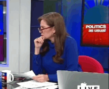 a woman wearing glasses is sitting at a desk with a laptop in front of her and a screen that says politics as usual